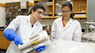 Two female international students wearing lab coats and goggles work on research at a lab at UMass Boston.
