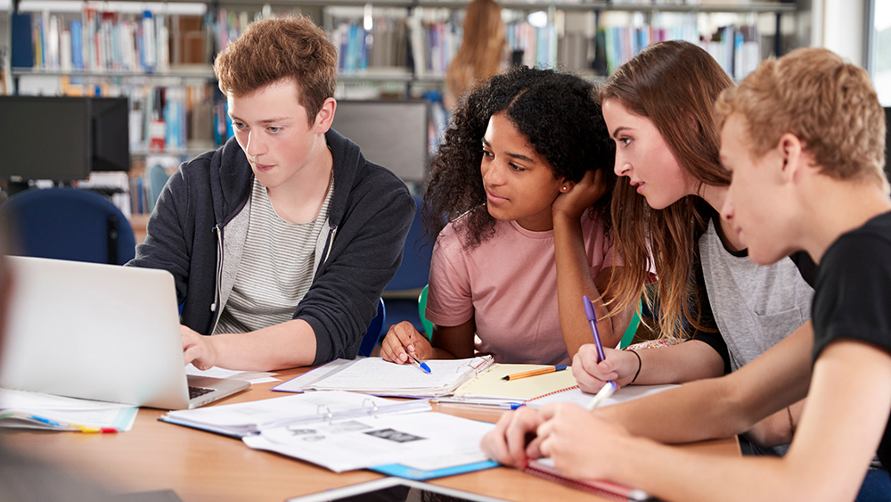 A group of four British students from the UK sit at a table around a laptop and work on applications for a study in USA visa for UK international students.