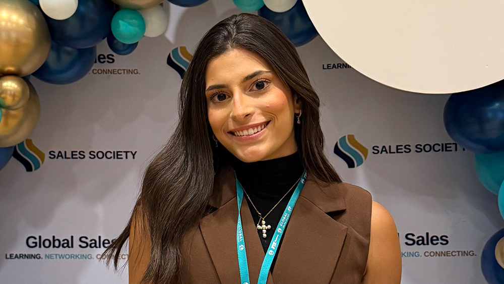 International student Ana is smiling at the camera; in the background is a backdrop at a professional event for the Sales Society.