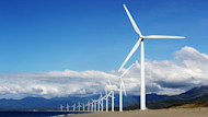 Wind turbines in a row line up before a mountain range and blue sky with white clouds