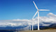 Wind turbines in a row line up before a mountain range and blue sky with white clouds