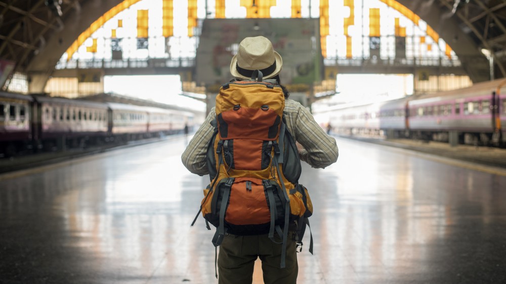 An international student in a hat and travel backpack facing away in an empty train station