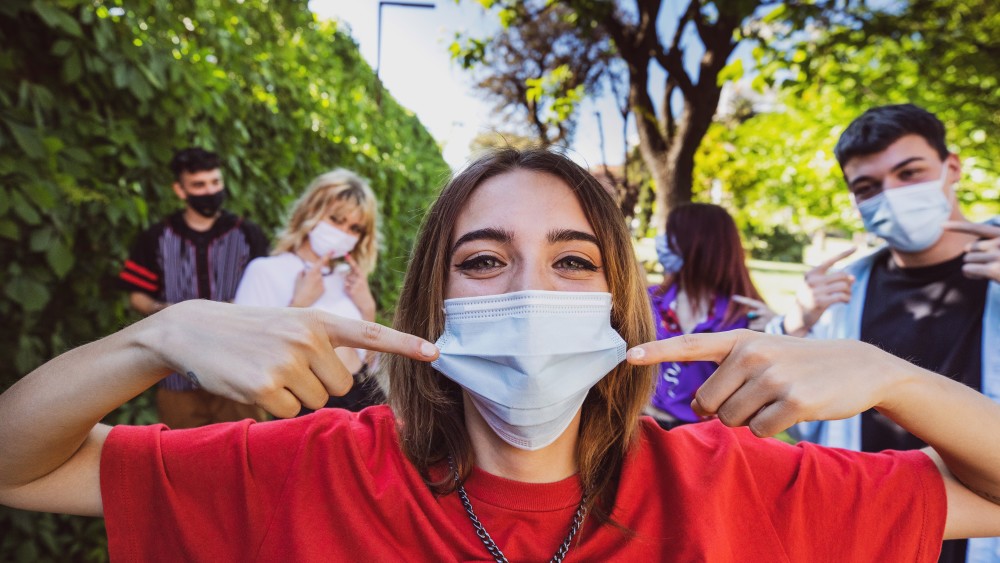 A female international student stands outside on a US university campus on a sunny day and points to the face mask she's wearing while two fellow masked students stand behind her