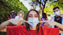 A female international student stands outside on a US university campus on a sunny day and points to the face mask she's wearing while two fellow masked students stand behind her