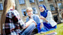 A blond male international student with headphones and a female international student wearing a blue burka sit outside across from a male student sitting cross-legged on the lawn of a college campus quad. 