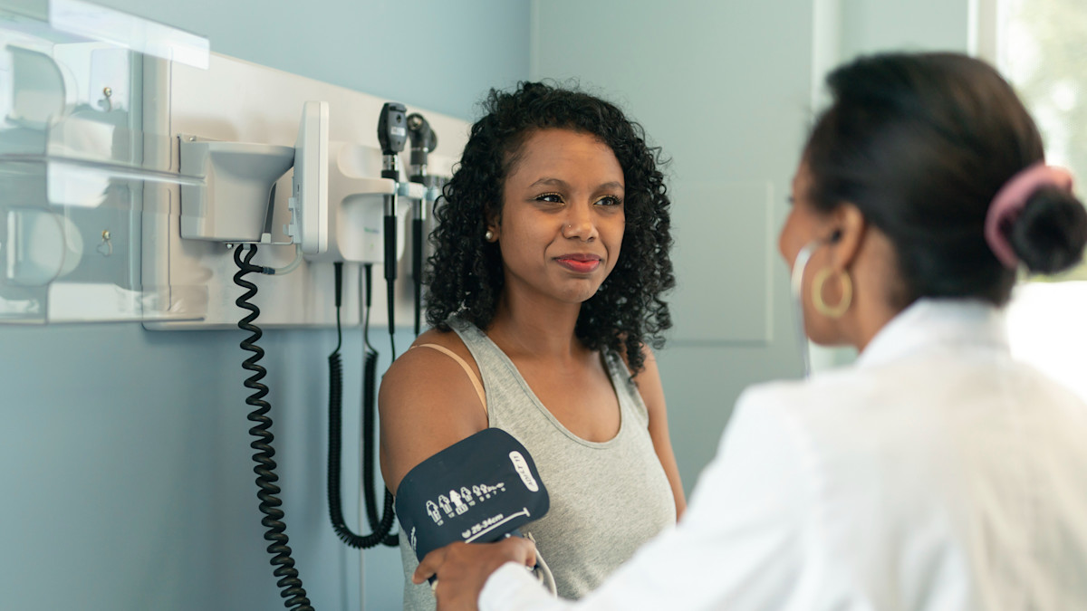 A woman has her blood pressure checked by a health worker wearing a white coat.