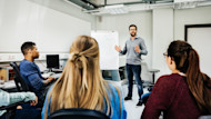 A professor stands next to an easel in front of two international students seated in a US university classroom. The students are learning what is a minor in planning their majors and minors for their degree programs. 