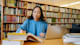 A female international student sits at a table with a stack of books in her US university library, doing research for an upcoming term paper.