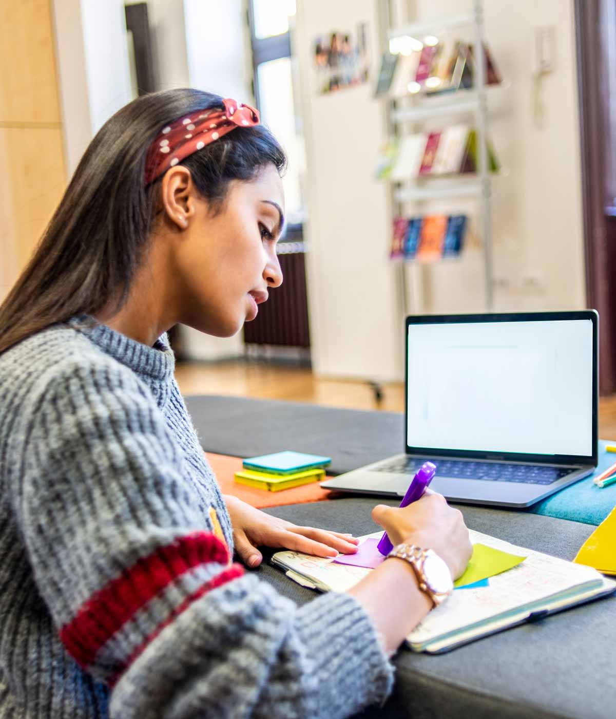 Female international student in a gray woolen sweater and red headband writing sticky notes with her laptop open at a table