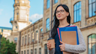 A female international student holds notebooks and a cup of coffee in front of a university hall on campus in the USA