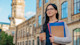 A female international student holds notebooks and a cup of coffee in front of a university hall on campus in the USA