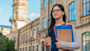 A female international student holds notebooks and a cup of coffee in front of a university hall on campus in the USA