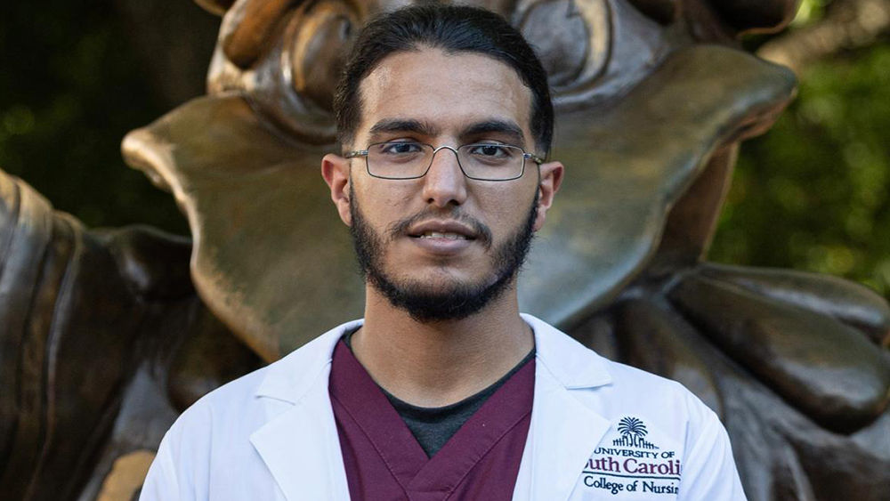 Ali, an international student from Saudi Arabia studying nursing at the University of South Carolina, stands in front of a statue of the school mascot on campus wearing a lab coat.