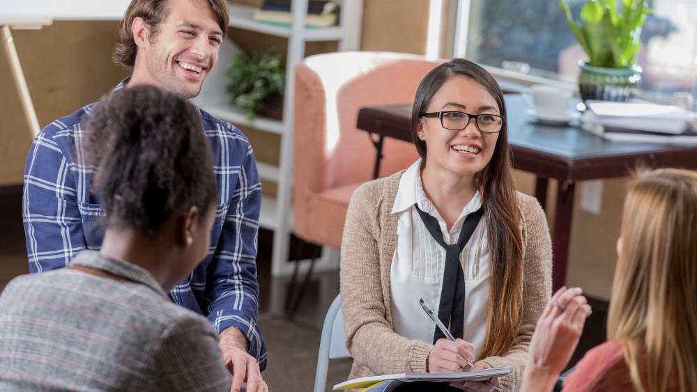 A female international student from Asia wearing glasses, a cardigan, and a tie sits across from another female student and takes notes in a notebook