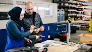 A female international student wearing a hijab stands in front of a table with an electrical drill, tools, and papers and works on a hands-on mechanical engineering project with her professor at a US university