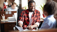 A smiling male international student in a red plaid shirt sits across from a female interviewer during a student visa interview. 