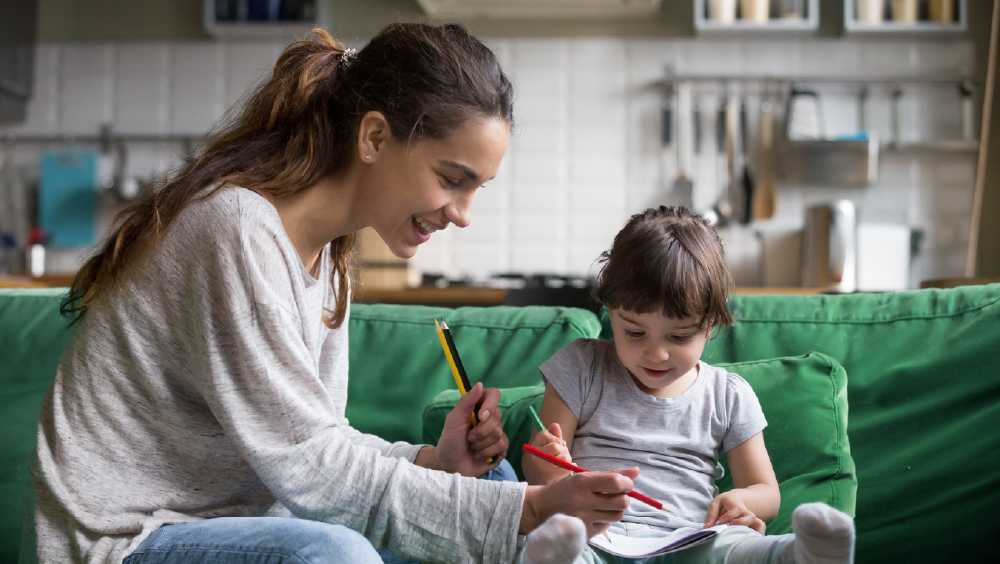A female international student sits on a sofa with a young child during her babysitting job. 