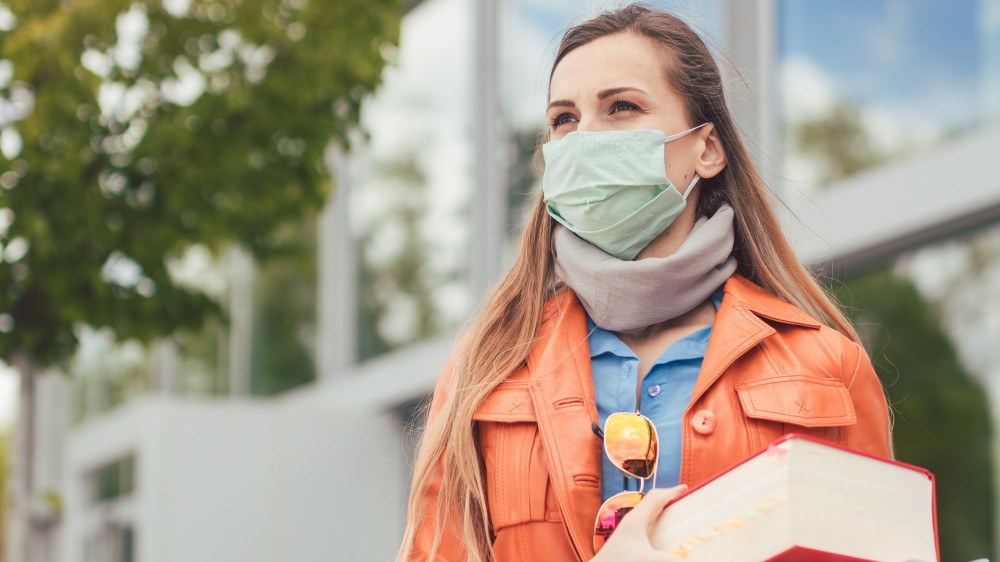 A female international college student holding textbooks and wearing a face mask stands outside on a college campus