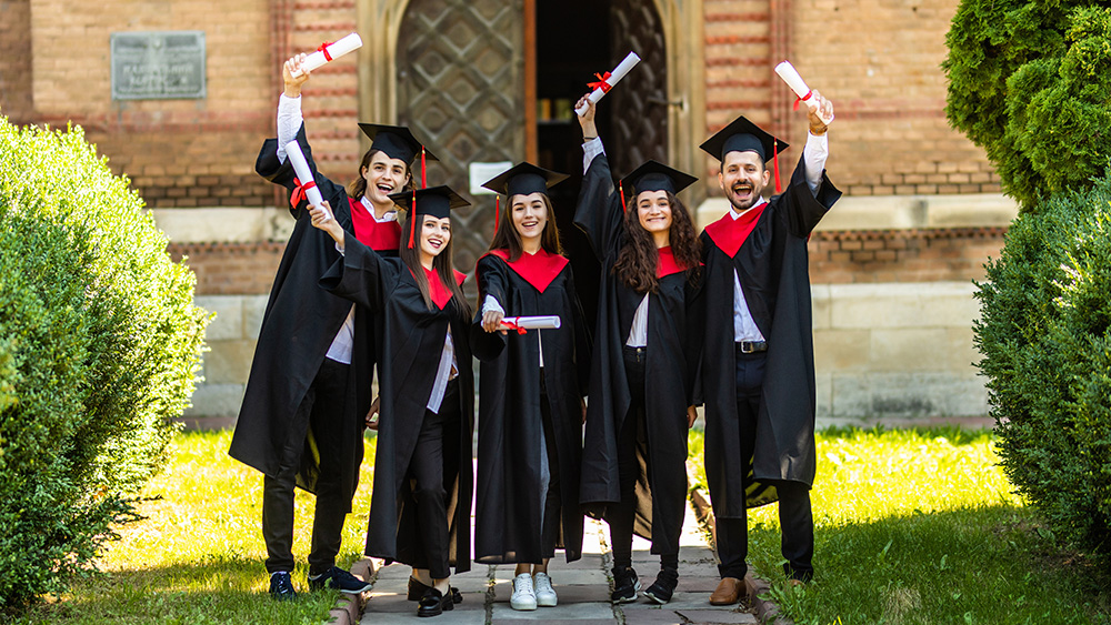 A group of international students in caps and gowns stand on their US university campus and hold up their diplomas after receiving their associate degrees.