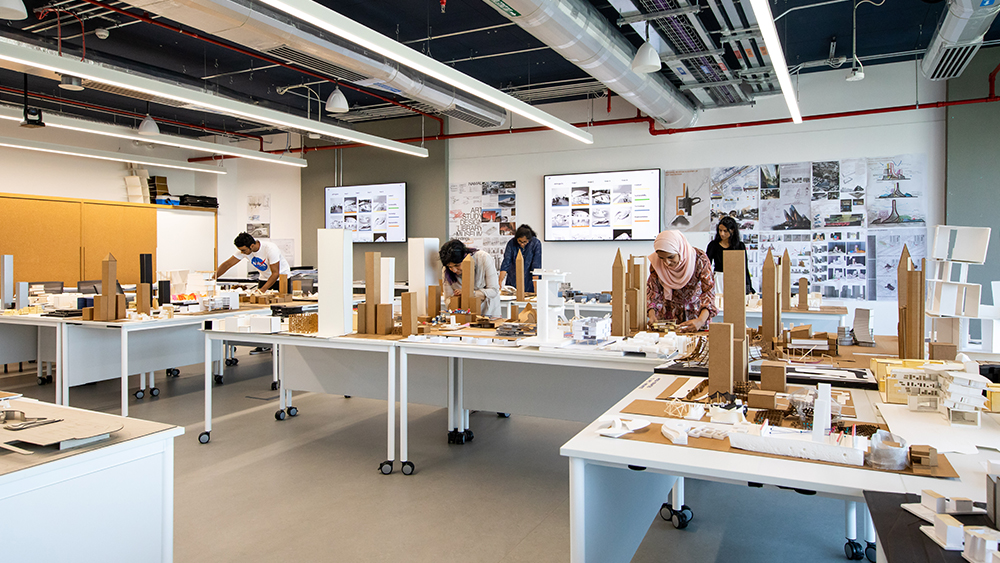 International students studying architectural engineering at Heriot-Watt Universit Dubai stand over model replicas of a city in their architecture classroom.