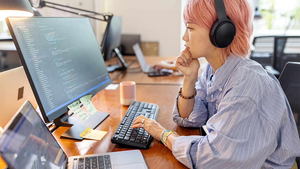 A female data analytics international student with short pink hair sits in front of two computer screens and analyzes data.