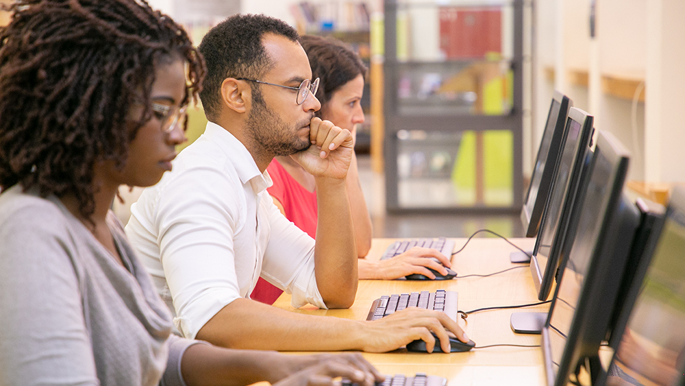 Three international students sit in a row at a table of workstations in the library at their accredited US university.