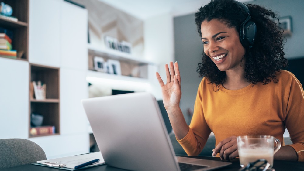 A female international student wearing headphones waves toward her laptop screen while taking an online orientation for American Collegiate Live