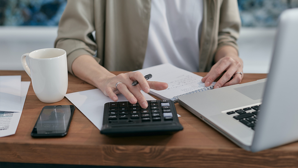 This photo shows the hands of a future accountant working on accounting spreadsheets with a calculator, laptop, and notebook as she does homework for her accounting courses at a US university. 