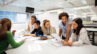 Five employees wearing company lanyards seated at a table, collaborating on work with a whiteboard in the background.