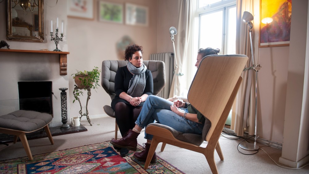 An international student sits on a wooden chair in a softly lit room across from a therapist on an ergonomic armchair during a mental health talk therapy session