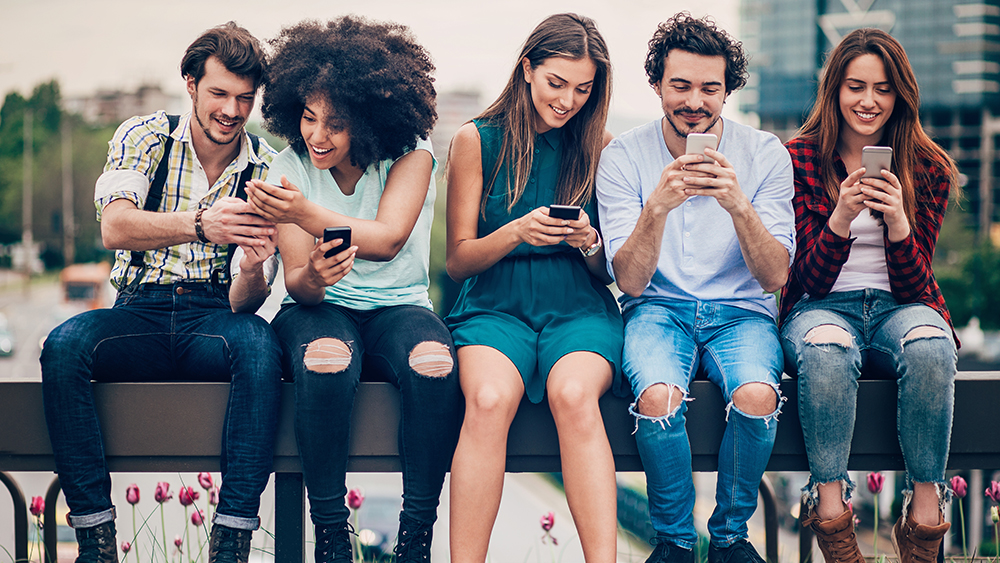 Five students sitting on a railing using English apps on their smartphones to practice their English language skills.