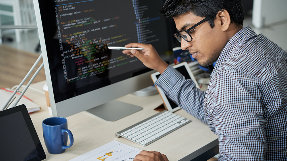 A male international student from South Asia sits at a computer screen showing code and works on an assignment for an IT course at his US university.