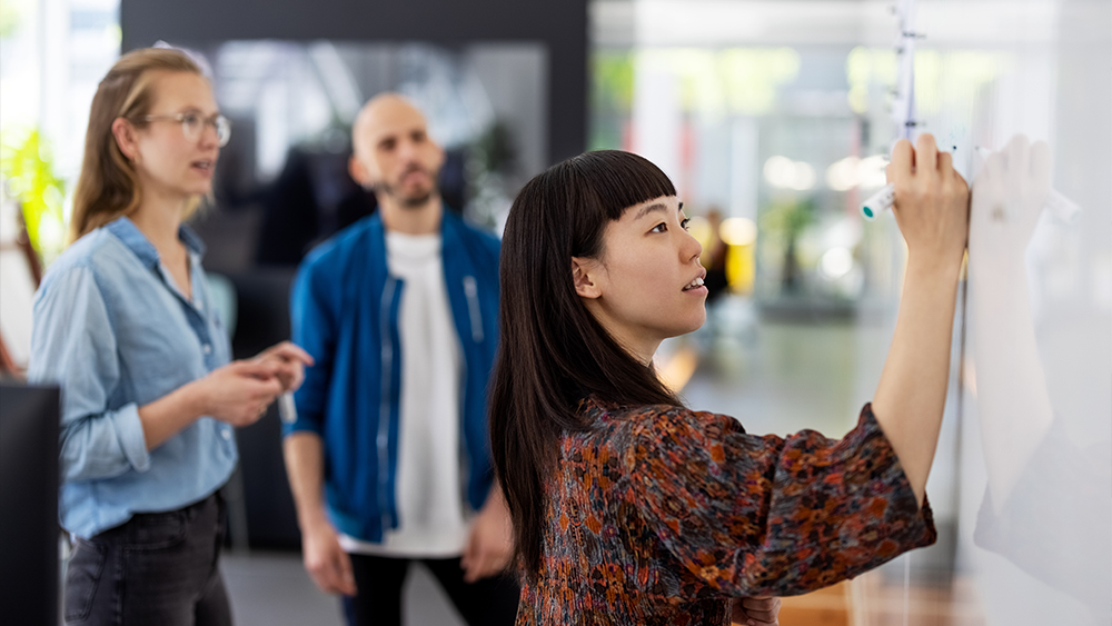 A female international student studying graphic design at a US university stands at a white board next to two professors and sketches out ideas for graphic design courses and web development projects.