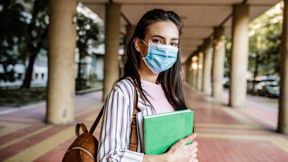 A female international student stands in an open-air hallway wearing a face mask and holding a textbook