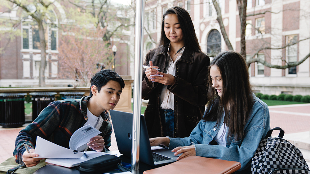 Three international students gather around a table on their US university quad and study together with books, a laptop, and a smartphone