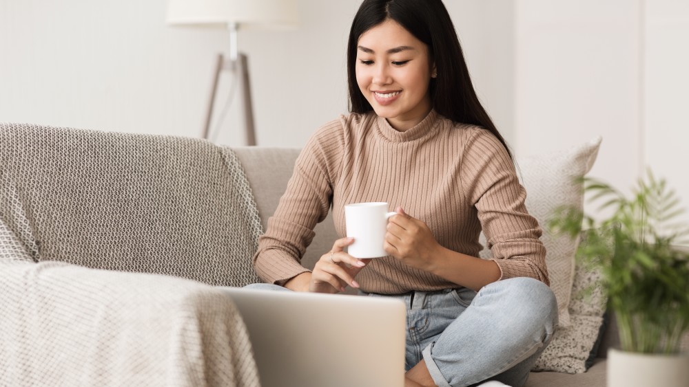 A woman sits cross-legged on her couch with a cup of coffee working on a laptop