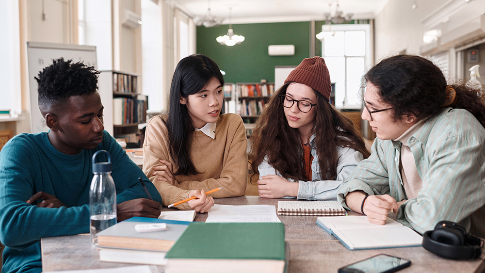 A group of four international students attending graduate school at a US university sit side by side at a table in a classroom and work on an academic project together.