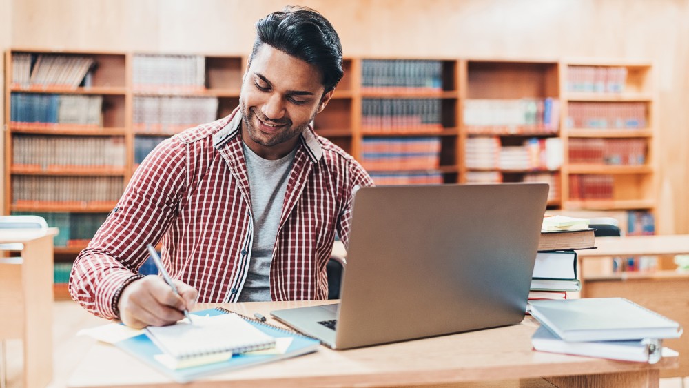 A male international student sits in front of a laptop in a university library and writes essay notes on a notepad.  