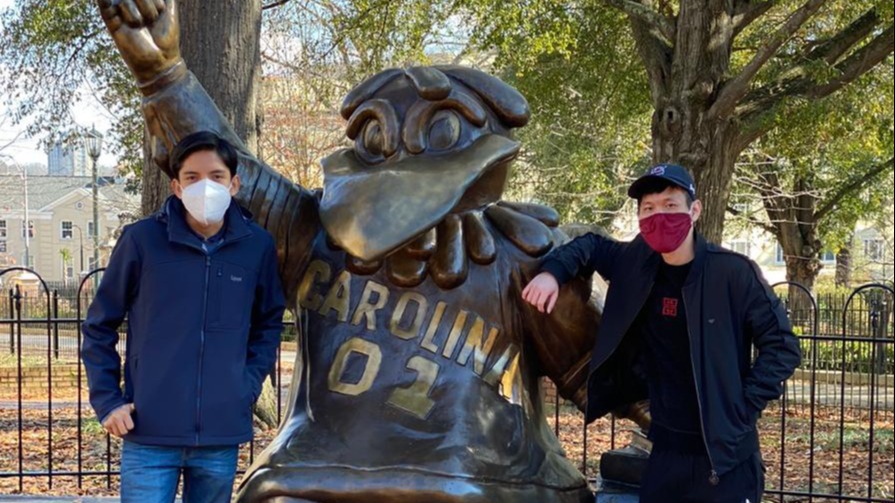Two international students at the University of South Carolina, a mentor and mentee, wear facemasks and stand next to a bronze statue of the South Carolina gamecock mascot.
