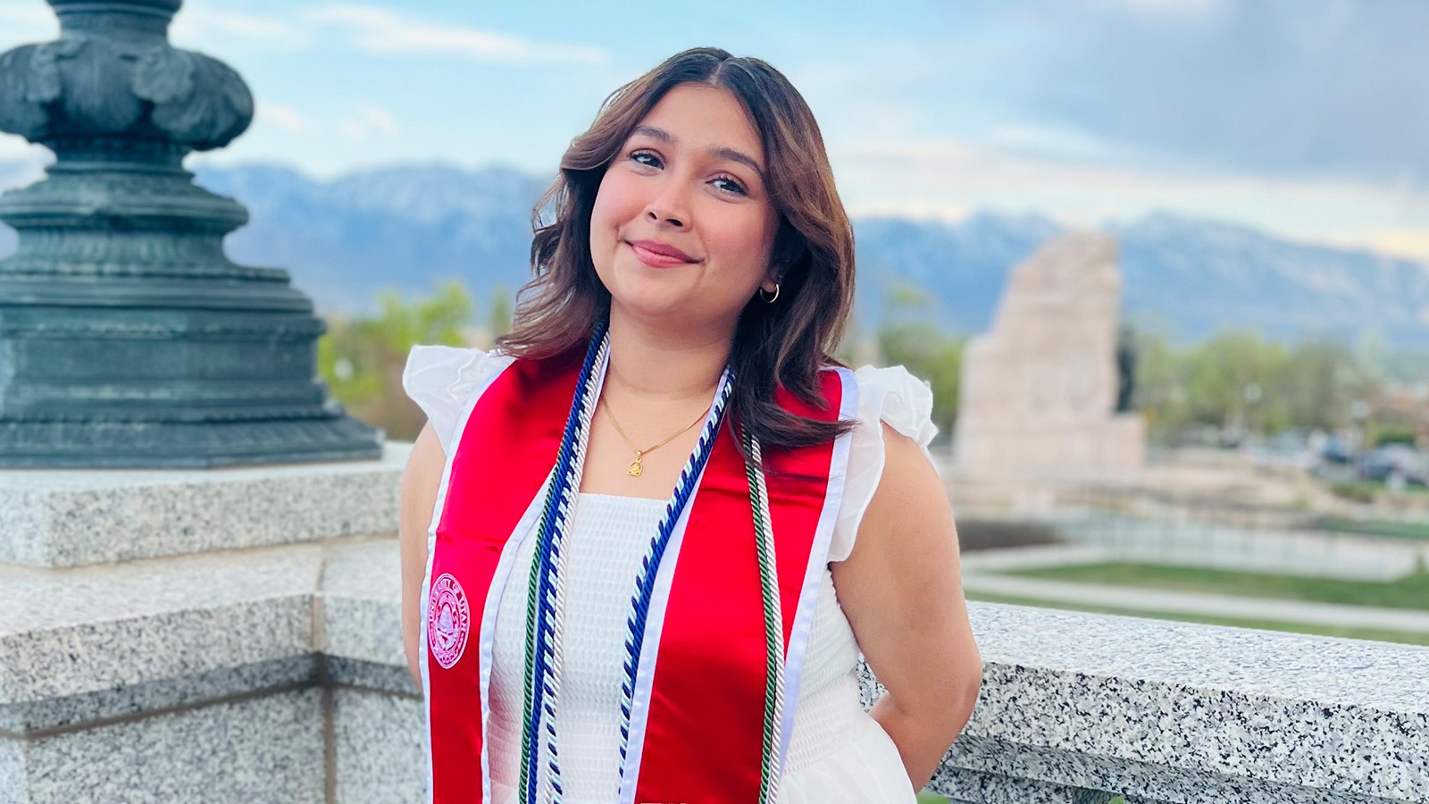 Aastha, a graduate of the University of Utah, stands on a balcony overlooking campus in her graduation ribbons and stole and smiles for the camera