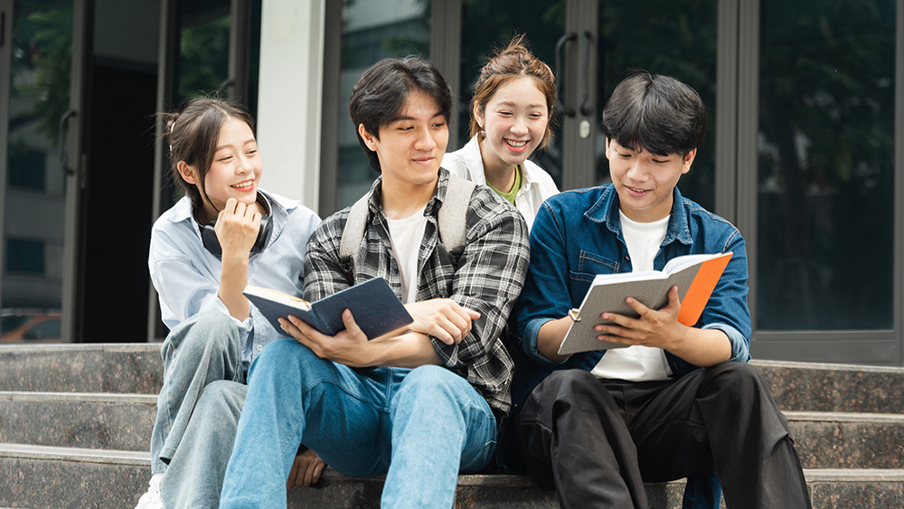 Four international students from Indonesia sit on the steps of their US university library and work on an assignment together.