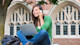 A female international student sits outdoors on a US college campus with a laptop on her lap while talking on a cell phone