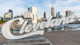 The Cleveland Script sign sculpture at Edgewater Park is in the foreground of this image, the Cleveland skyline and Cuyahoga River is in the background.