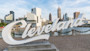 The Cleveland Script sign sculpture at Edgewater Park is in the foreground of this image, the Cleveland skyline and Cuyahoga River is in the background.