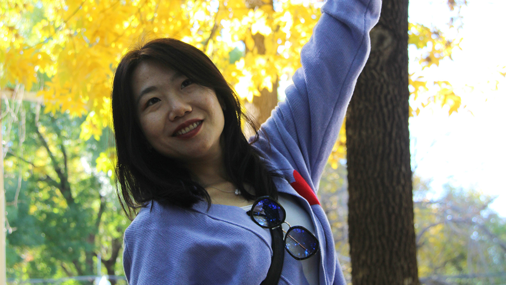 Alicia Liu, student success coordinator for Shorelight, stands in front of a tree with yellow fall foliage leaves and smiles for the camera.