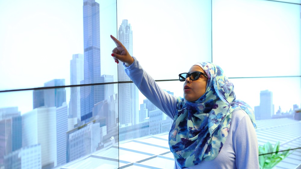 A UIC female international student wearing a hijab and glasses stands in front of a wall of windows overlooking the Chicago skyline and points upward