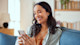 Smiling woman sitting on a couch, looking at her smartphone in a bright living room.