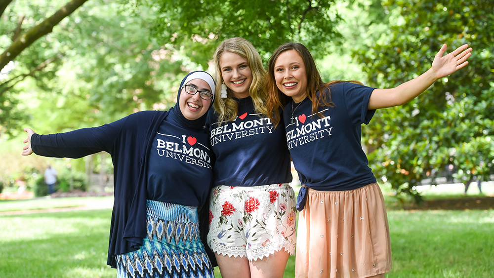 Three female international students wearing I heart Belmont University stand outside on Belmont University's campus and smile for the camera