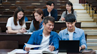 Two students sitting a lecture hall while looking at a laptop, with three other students seated behind them.