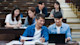 Two students sitting a lecture hall while looking at a laptop, with three other students seated behind them.